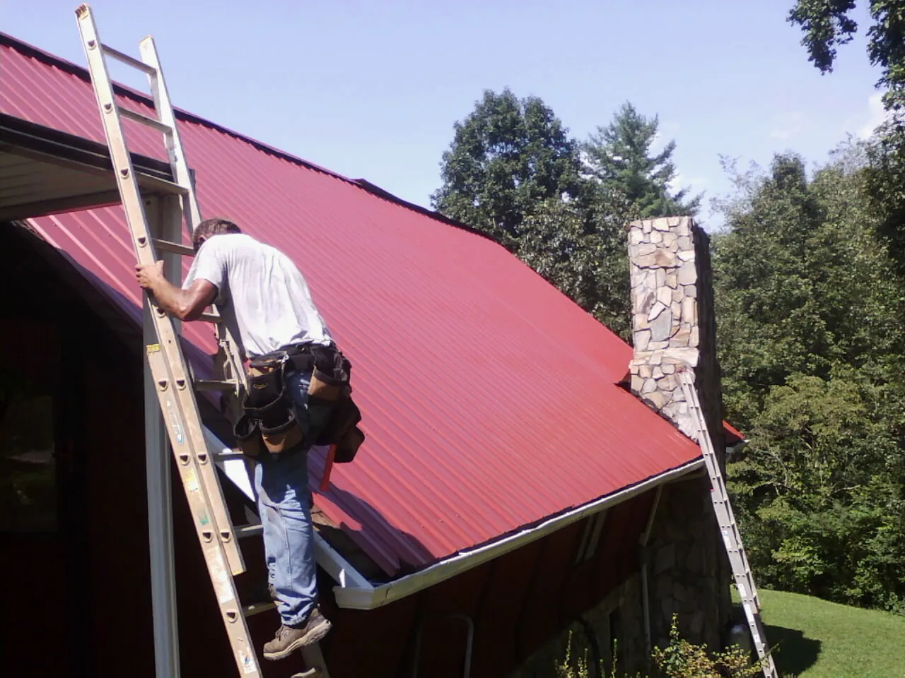 Roofer on a ladder beside a red metal roof and stone chimney — Soffit & Fascia Repair by Roofing Brothers in West Bridgewater